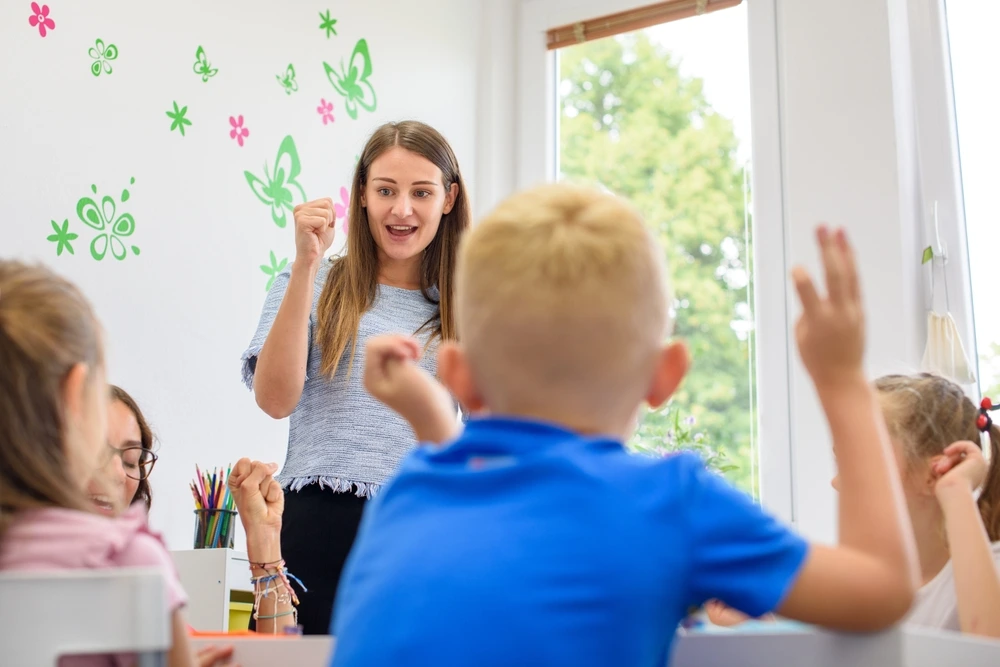 A female YBL Behaviour Support member in a classroom, engaging with several children who are raising their hands