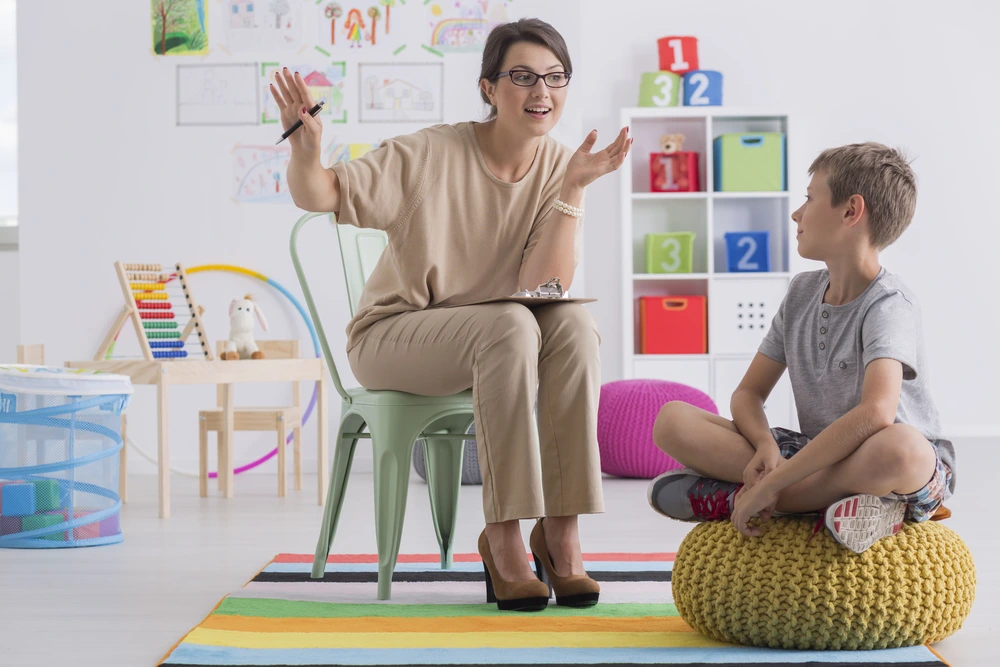 A YBL Behaviour Support team member sits on a chair, talking with a boy who is seated on a yellow cushion